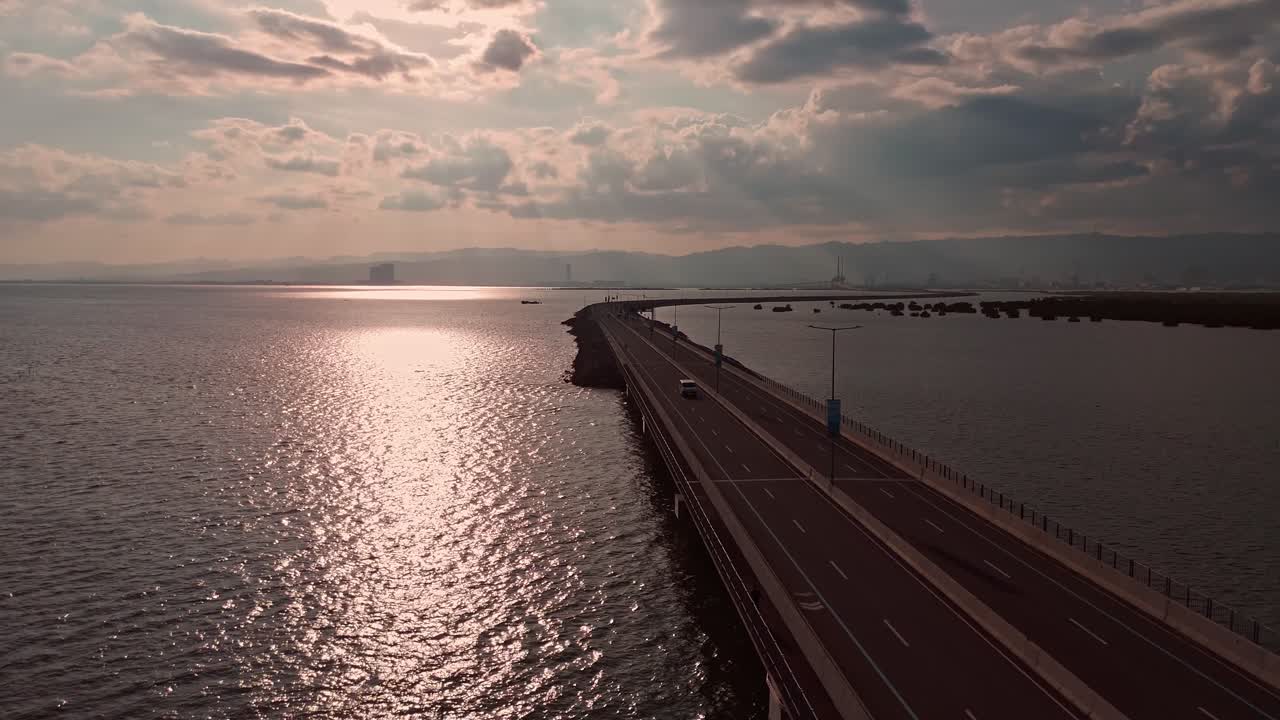 Stunning drone footage of a coastal highway bridge over water at sunset with shimmering reflections and mountain backdrop, ideal for travel, cinematic, and infrastructure projects