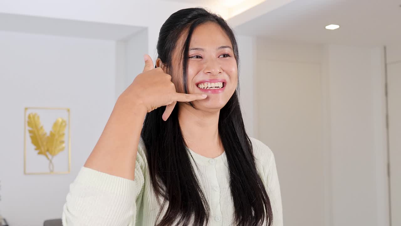 A woman in a white room smiles and gestures a phone call with her hand, conveying a cheerful mood