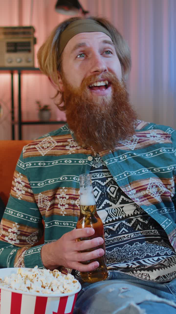 Mand sidder på sofaen, spiser popcorn og ser en spændende tv-serie eller sportskamp online derhjemme.