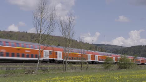 an orange train speeds through a scenic countryside with wind turbines and trees in the background