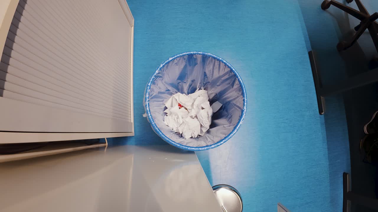 Overhead office shot of a wastebasket as a crumpled sheet of paper is tossed in, capturing a simple daily workplace moment. Ideal for concepts like rejected ideas, mistakes, cleanup, productivity, and