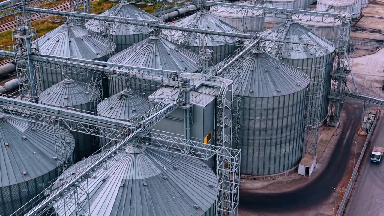 Vista de grandes silos de armazenamento para grãos em uma instalação industrial. Silos se erguem em uma área industrial. Equipamentos conectam as estruturas. É dia e o céu está limpo.