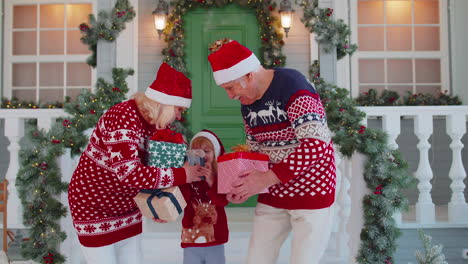 Senior-grandparents-with-grandchild-girl-kid-exhcanging-gifts-near-Christmas-house-celebrating-Xmas