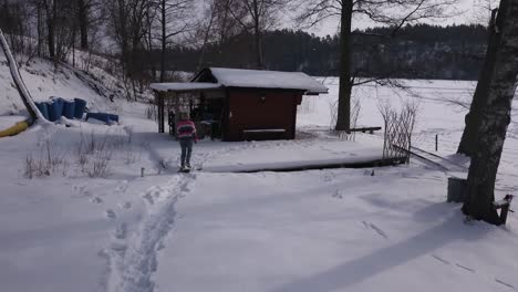 Aerial-follow-shot-of-young-blonde-girl-walking-toward-small-cabin-in-forest-on-the-edge-of-a-big-frozen-lake