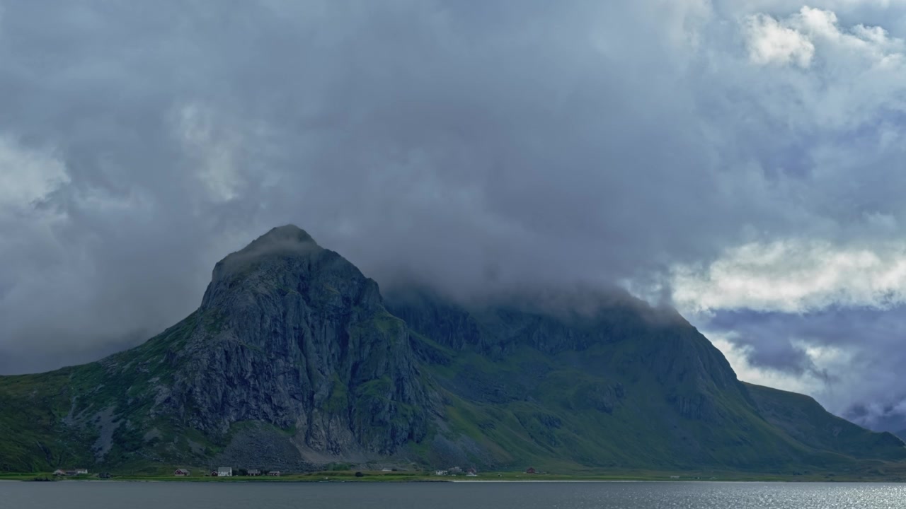 Cima di montagna avvolta dalle nuvole (Time-Lapse). Case su un'isola ai piedi di una maestosa montagna sul mare in una nuvolosa giornata estiva polare. Flakstad, Isole Lofoten, Norvegia settentrionale.