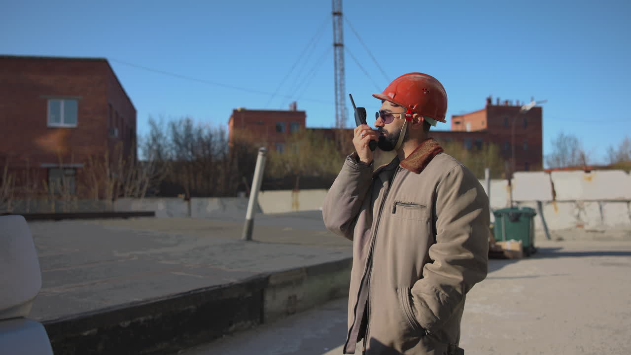 Supervisore caucasico barbuto in cantiere con casco rosso e occhiali da sole che usa un walkie-talkie e ispeziona.