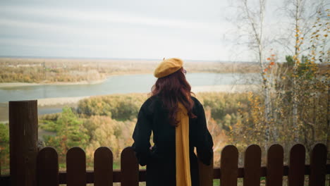 back view of a woman with red wavy hair in a black coat, walking towards a wooden fence, resting her hand on it while enjoying the scenic view of a river and dry foliage