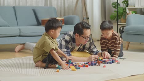 full body of asian father and sons playing the construction set colorful plastic toy brick at home. the kids assemble plastic building blocks sitting on a mat, a father lying on the floor talking to his sons