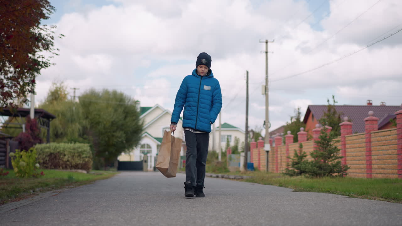 파란색 재과 검은색 베니를 입은 어린 소년은 쇼핑에서 돌아오는 종이 가방을 들고 교외 거리를 고 있으며, 주거 주택과 가을 나무가 길을 고 있습니다.