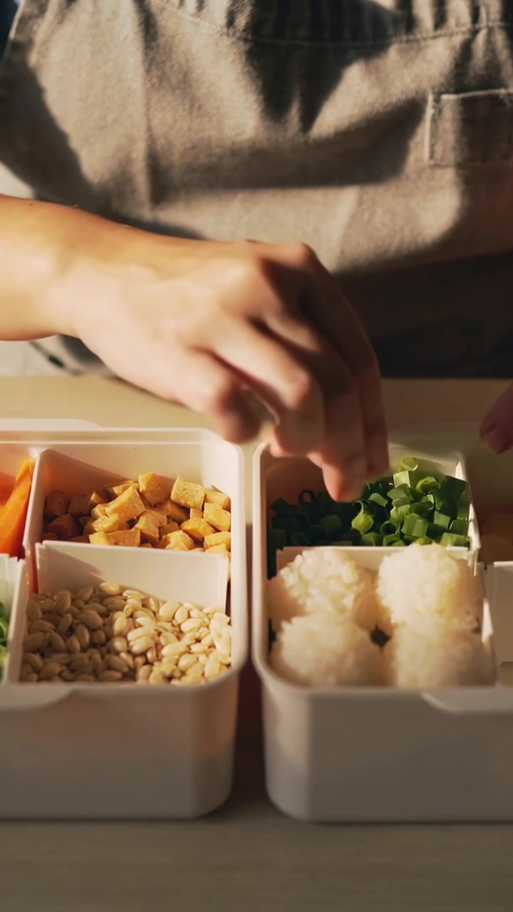 Vertical video: Reaching cook in gray apron placing onions into bento at table for rice ball lunch