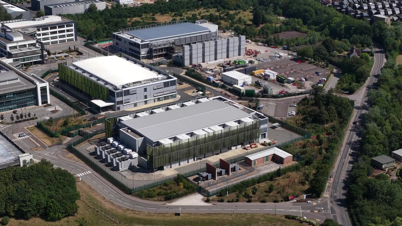 Vidstrakt luftfoto, der langsomt cirkulerer moderne erhvervsparkbygninger, og fremviser den moderne arkitektur på et stort teknologicampus og datacenterkompleks omgivet af grønne træer