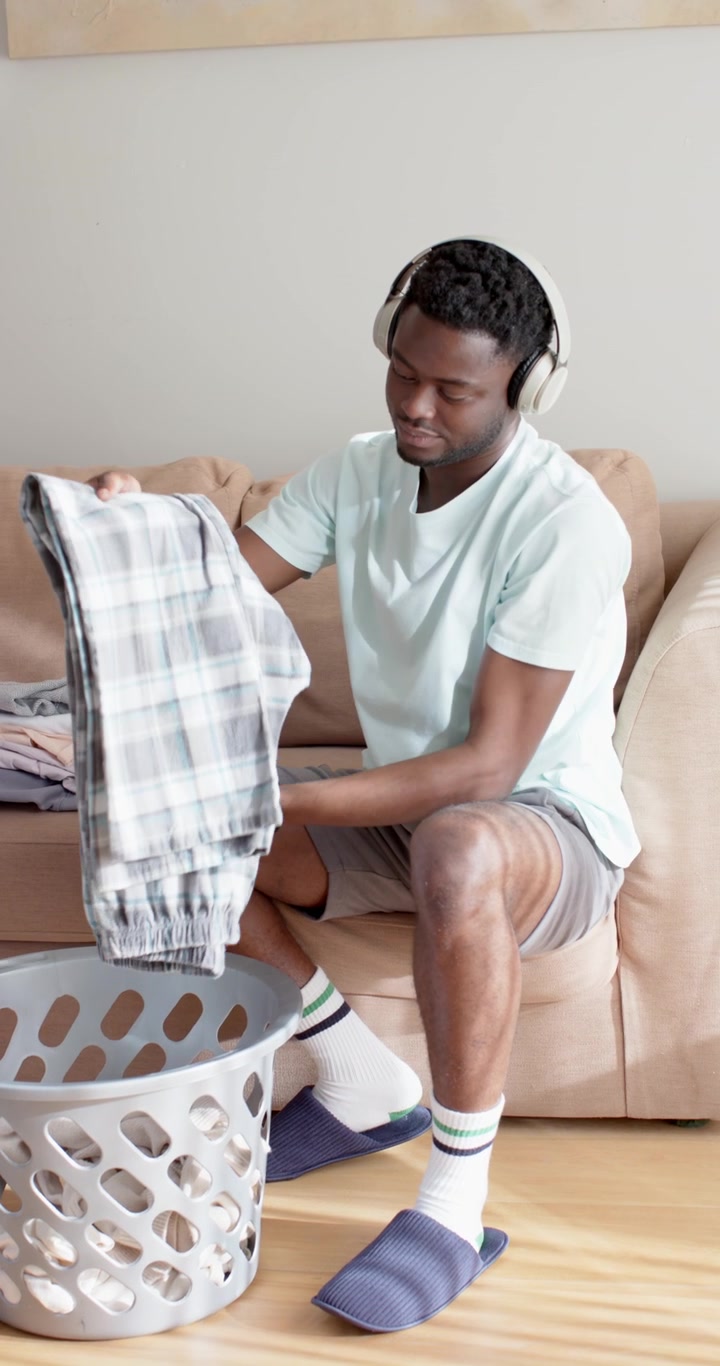 Vertical video: African American man folding laundry on sofa, reaching into basket to stack clothes