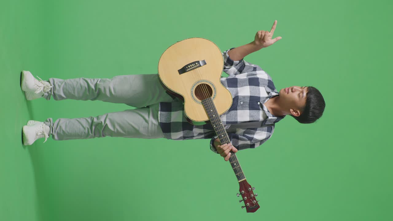 Figura intera di adolescente asiatico con la chitarra che sorride e indica di lato su sfondo verde.