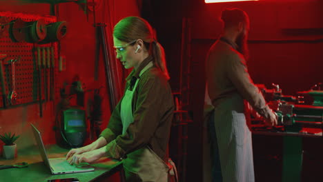 retrato de mujer joven en delantal y gafas usando una laptop en un taller de metal mientras su compañero de trabajo está usando una máquina de metal