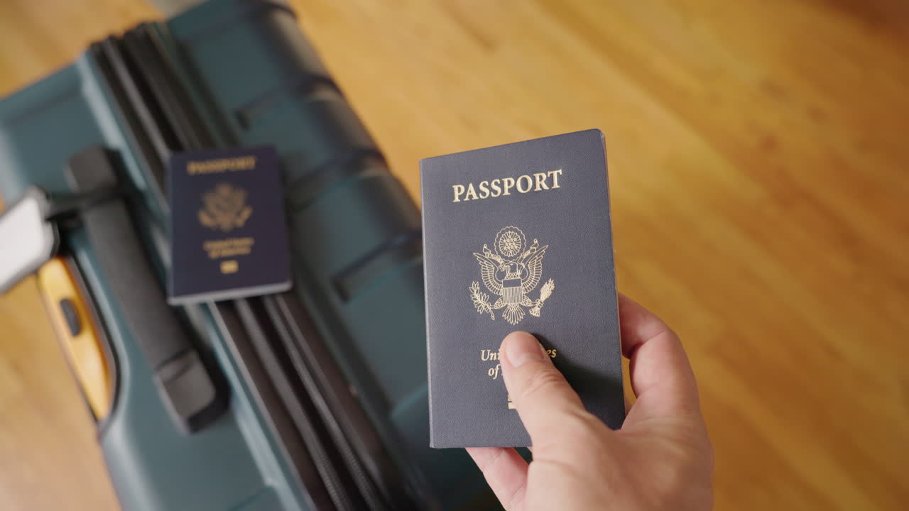 Mano che regge un passaporto USA con una valigia sullo sfondo.