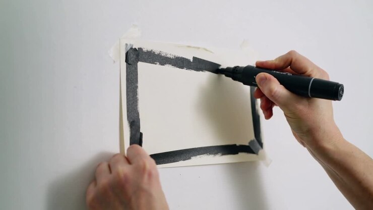 Drawing hands finishing black border on taped sheet on wall, using marker and tape, short sleeves