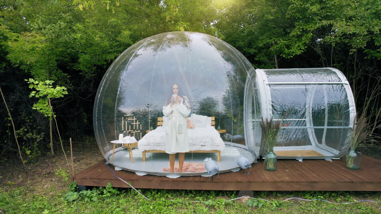 Tenda a bolla trasparente per il glamping, foresta lussureggiante circostante e interni. Una donna si alza dal letto con una tazza e guarda fuori