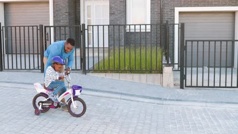 Feliz-Padre-Afroamericano-Enseñando-A-Una-Pequeña-Niña-Bonita-Con-Casco-Montando-En-Bicicleta-En-La-Calle-En-Las-Afueras