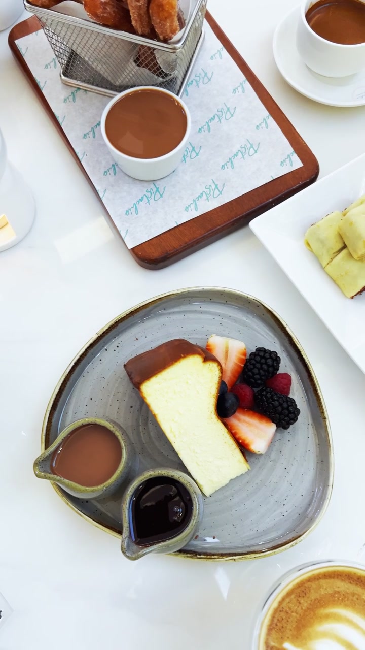 Top-down view of a bright cafe table spread featuring a slice of cheesecake with fresh berries, sweet rolled crepes, sliced bananas, pineapple, and a cup of latte art coffee during daylight.