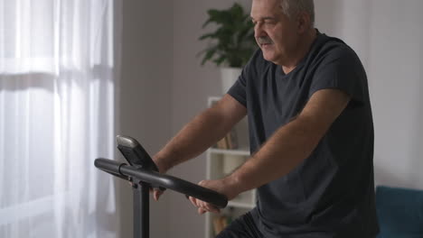 Un-Hombre-De-Mediana-Edad-Está-Entrenando-Solo-En-Casa-Usando-Una-Bicicleta-Estática-Moderna-Para-Hacer-Ejercicio-Preocupándose-Por-La-Salud-Y-Manteniéndose-En-Forma-En-La-Vejez