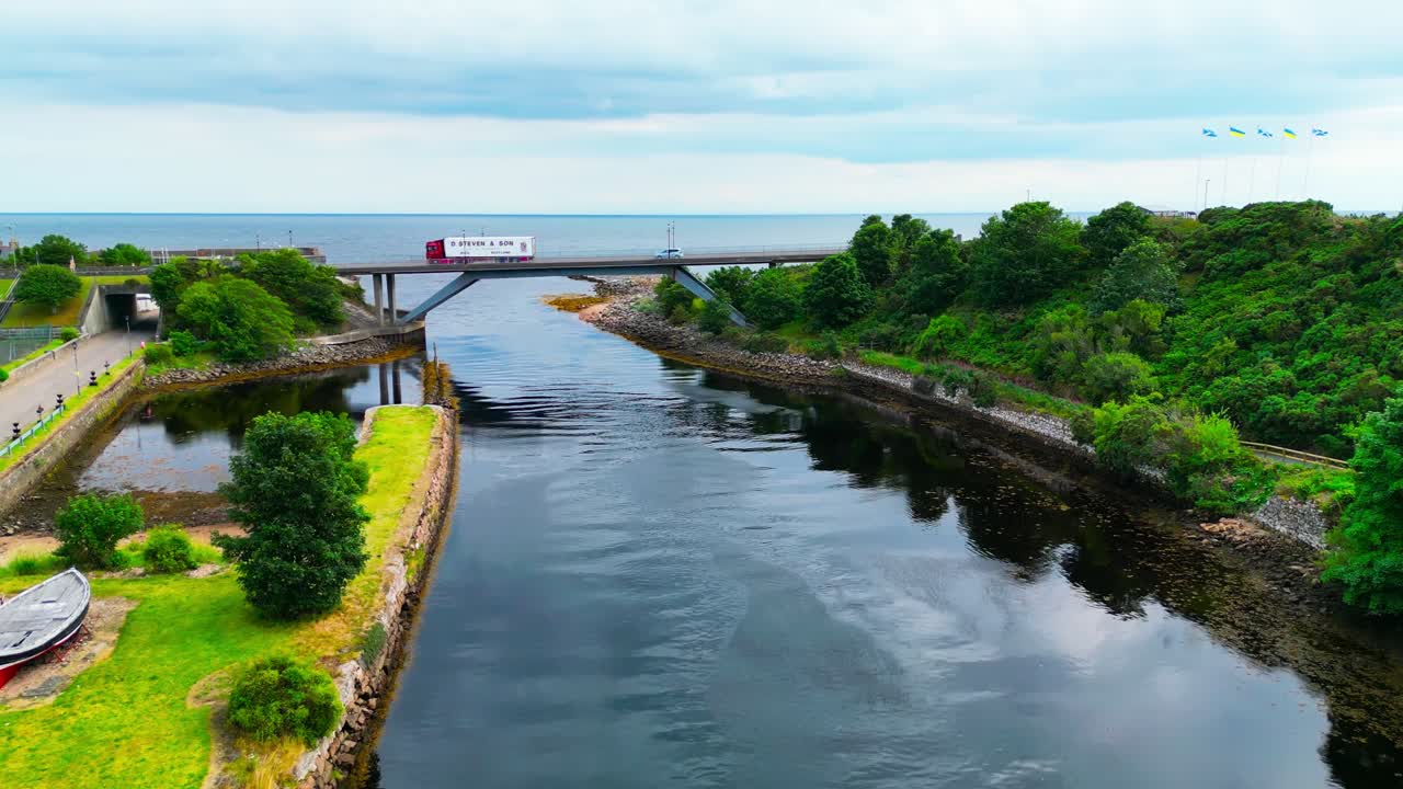 Ripresa cinematografica con drone che percorre il fiume Helmsdale sotto un ponte, salendo per rivelare il villaggio costiero di Helmsdale e il Mare del Nord, mostrando il paesaggio delle Highlands scozzesi.