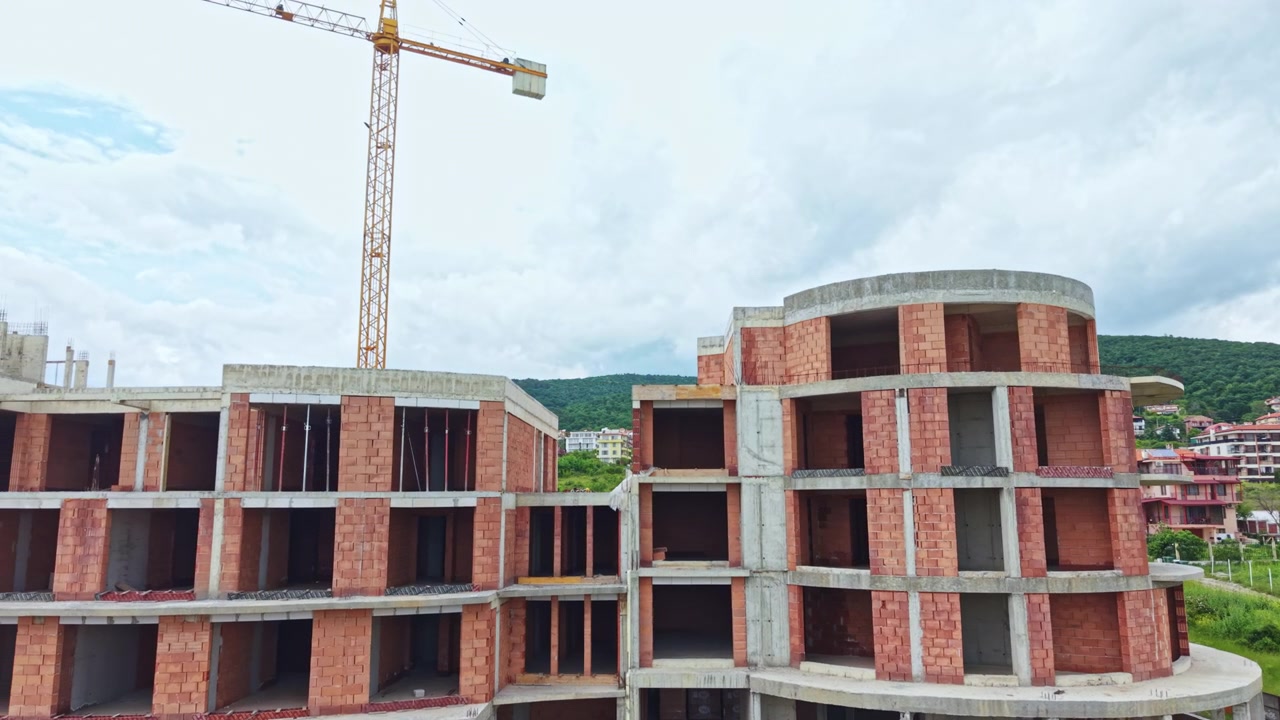 Desarrollo urbano en progreso con un pintoresco paisaje de fondo. La construcción de apartamentos de ladrillo sin terminar bajo una grúa destaca la constante expansión urbana y el paisaje natural.