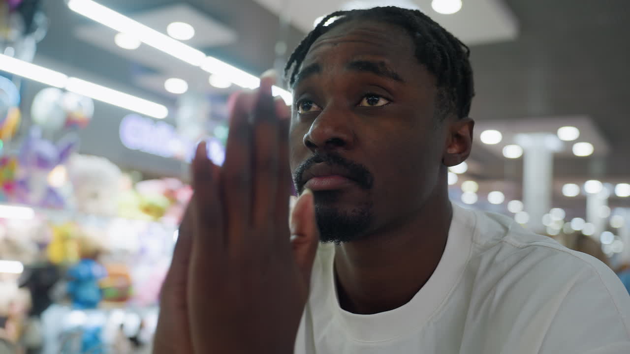 Tired young man seated in brightly lit colorful cafe looks ahead in silence while gently rubbing his palms together and touching his face with serious expression surrounded by bokeh background lights