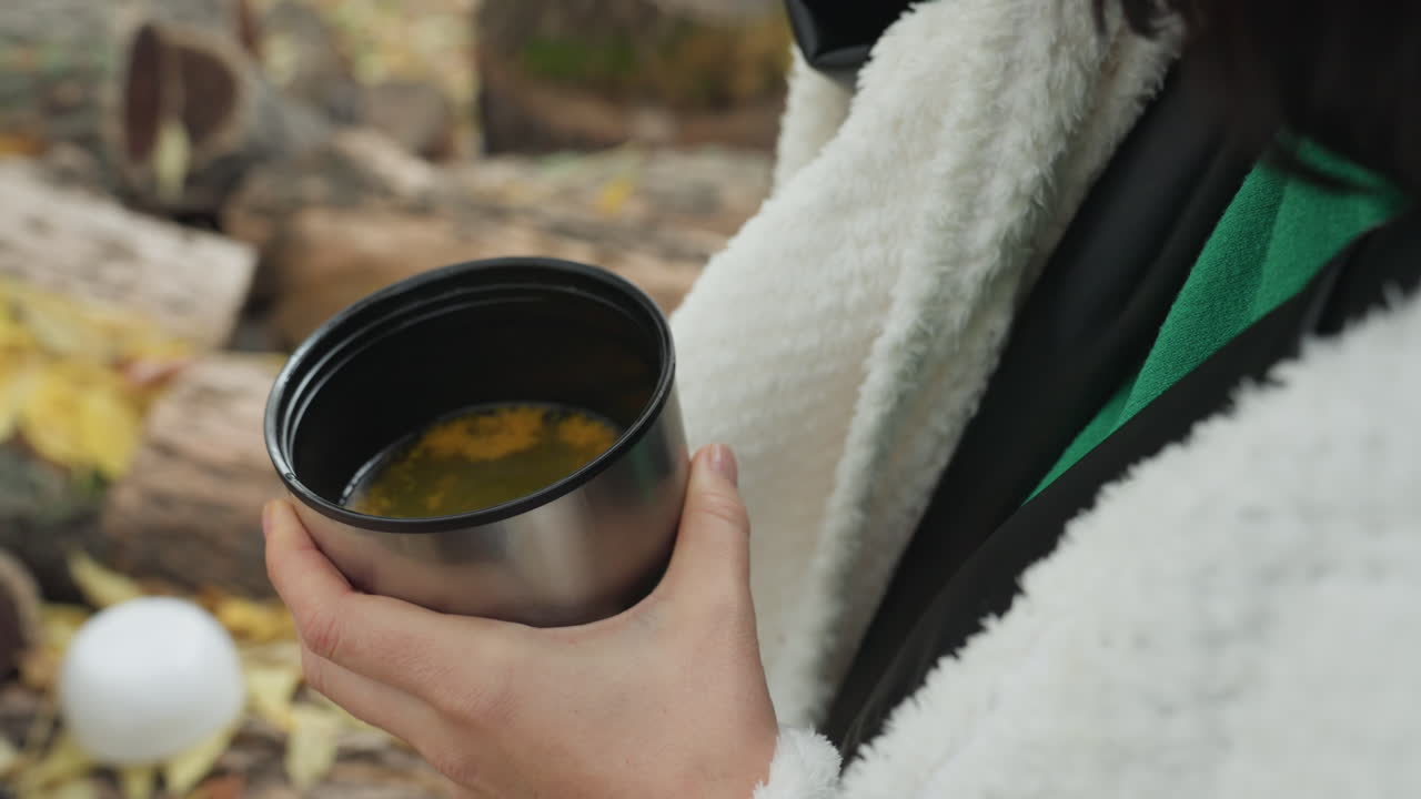 Primo piano all'aperto di una tazza di tè autunnale con mani che tengono una tazza in acciaio inossidabile su un tronco muschioso e foglie cadute, vapore che sale dall'infuso di erbe, comoda giacca in pile con un maglione verde che spunta, escursionista rilassato che si gode il momento