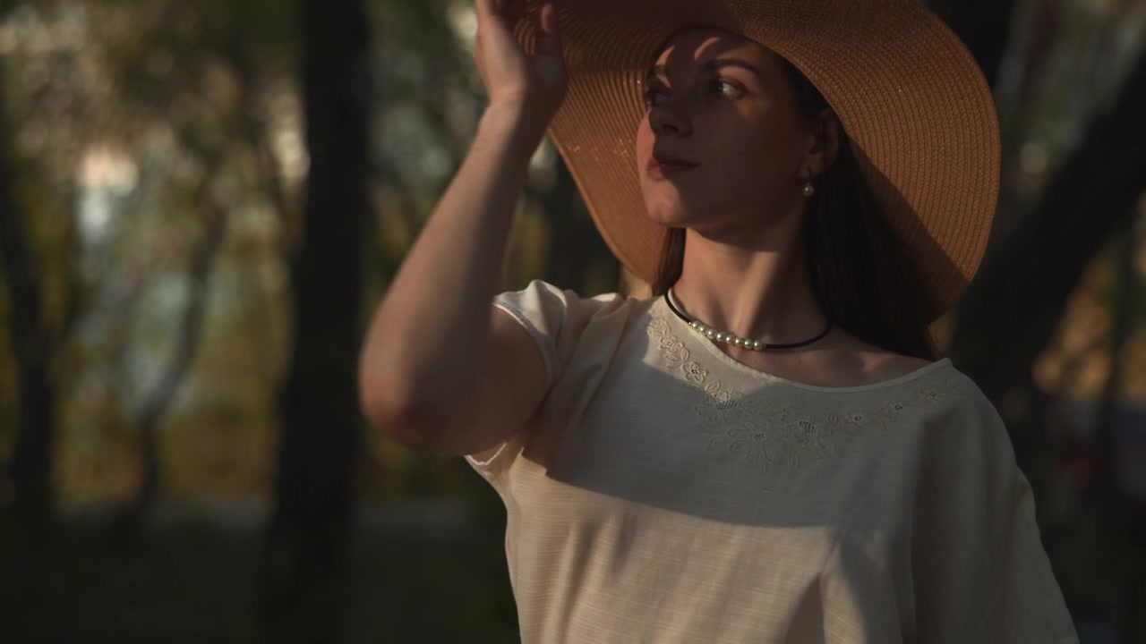 Una donna adulta alza il braccio in una radura boschiva con la calda luce del sole che filtra tra gli alberi, mentre una seconda donna con un cappello a tesa larga è parzialmente visibile.