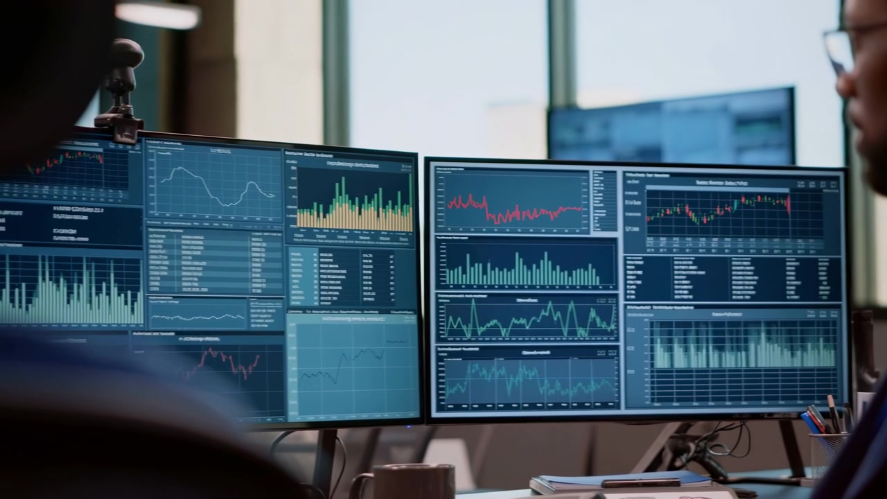 Multiple monitors show colorful charts and graphs as person works beside desk, with bright window light and office setting creating a professional technology focused workspace composition.