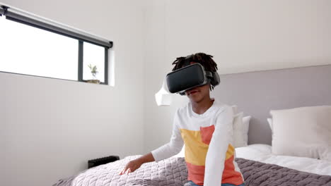 african american boy sitting on bed using vr headset at home, slow motion