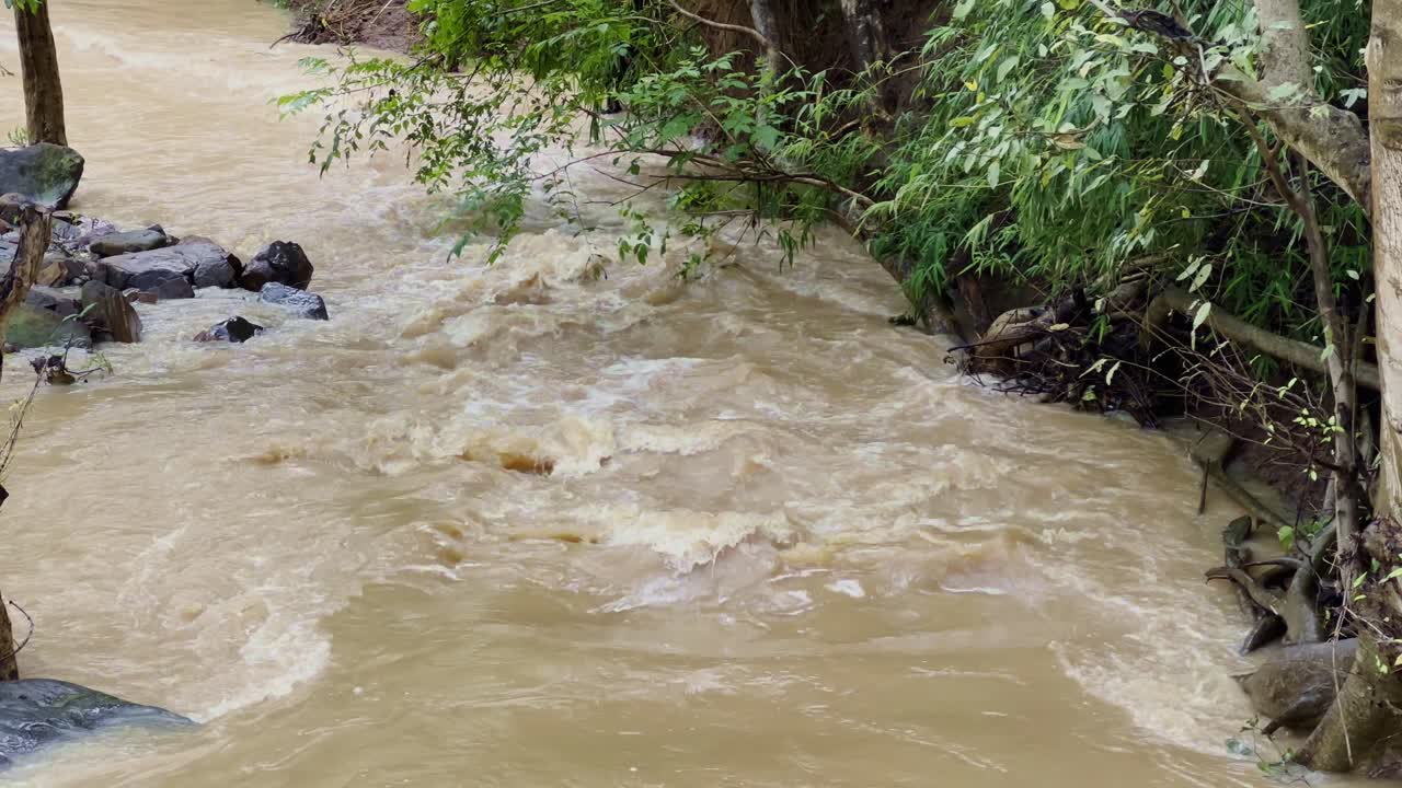 Fast-flowing muddy river water rushing downstream with rocks and trees along the banks, creating natural rapids