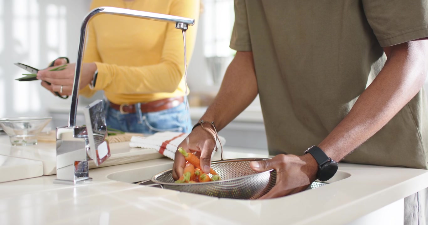 Couple préparant un repas dans la cuisine, l'homme ouvrant le robinet, rinçant des tomates dans une passoire pendant que la femme les prépare