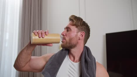 Confident-man-with-brown-hair-of-an-athletic-build-with-stubble-in-a-white-T-shirt-with-a-gray-towel-around-his-neck-drinks-water-from-a-sports-bottle-of-wooden-texture-after-doing-warm-up-and-exercise-exercises-at-home-in-a-modern-apartment