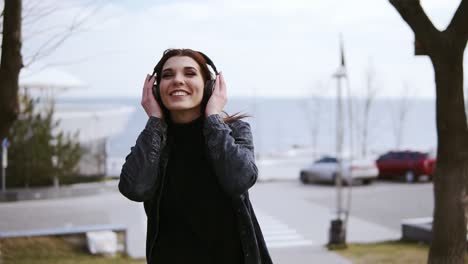 Attractive-young-brunette-girl-with-black-wireless-headphones-on-is-walking-somewhere,-listening-and-singing-to-music.-Enjoying-youth-and-free-time.-Outdoors-footage
