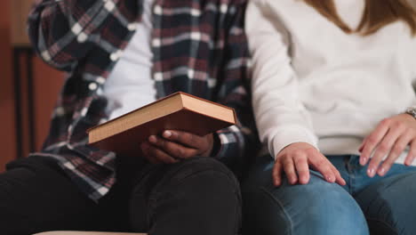 man with book sits next to woman in library closeup. black guy with textbook and white girlfriend in university. young couple studies together in college