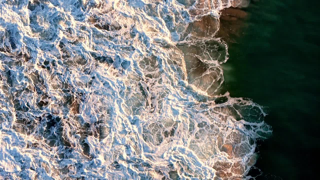 Aerial view of the foamy waves crashing against the shoreline, Sunlight casting a golden glow on the ocean's froth