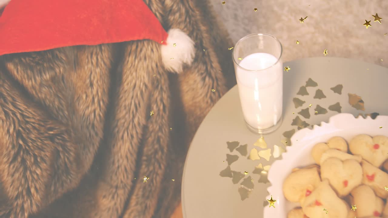 Confetti drifting over red Santa hat on throw, camera panning right revealing milk glass, cookies