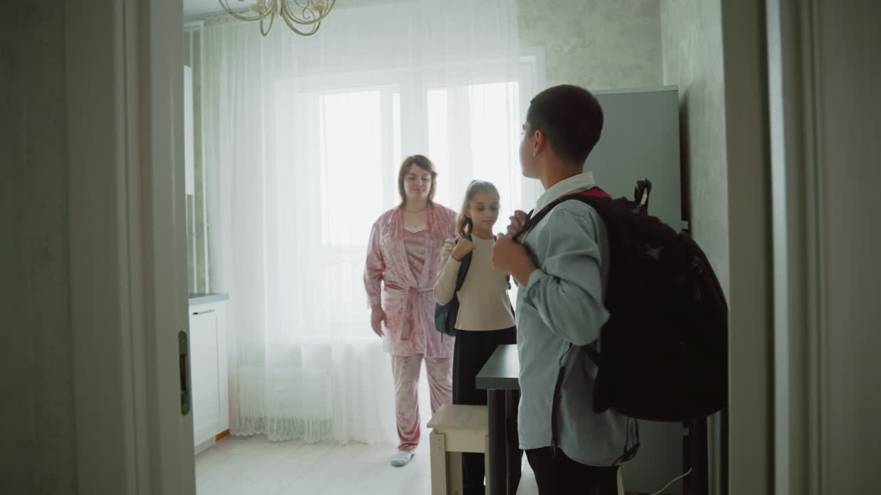 kids in casual wear organizing backpacks for school in bright modern kitchen as mother supervises morning preparation, boy adjusts straps while girl receives help, warm natural light fills cozy home