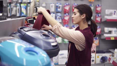 girl choosing vacuum cleaner in appliance store. coming up to a display row taking a dustbag out of vacuum cleaner. costumer examining domestic equipment.