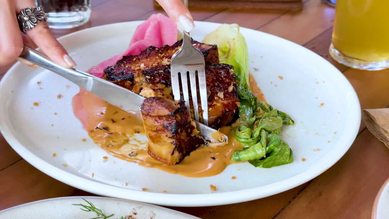 Hand uses knife and fork to slice grilled pork belly steak on a white plate with sauce, greens, and pickled vegetables in bright natural light
