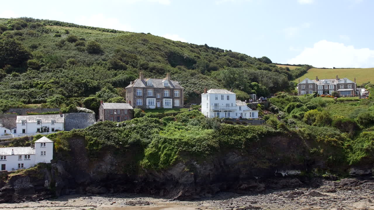 Houses perched along a cliff face overlook a rocky beach, backed by steep green hills and surrounded by dense vegetation and winding footpaths Port Isaac, Cornwall, England
