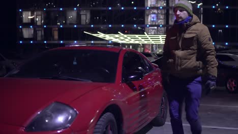 young man gets out of a red car at night at the parking and closes the door. front view.