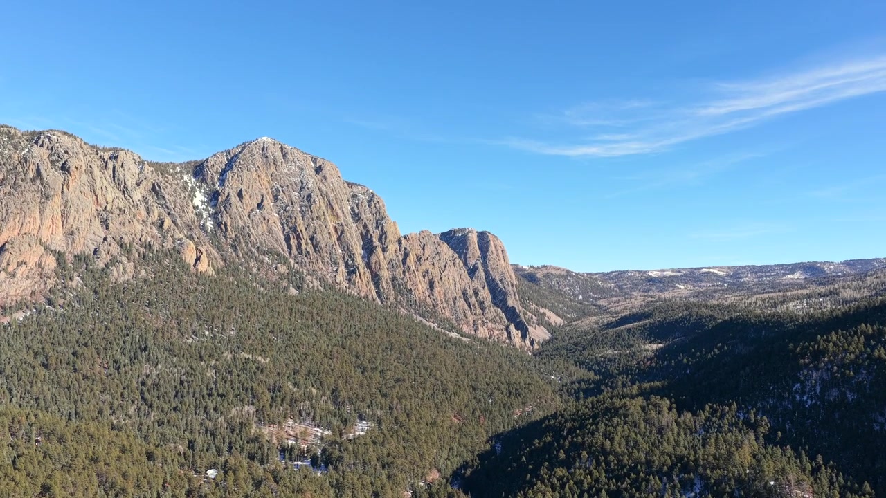 Filmato con drone che sorvola una fitta foresta sempreverde verso imponenti pareti rocciose, rivelando le scogliere di Brazos che si ergono su una valle di montagna con leggere chiazze di neve e un carattere alpino aspro