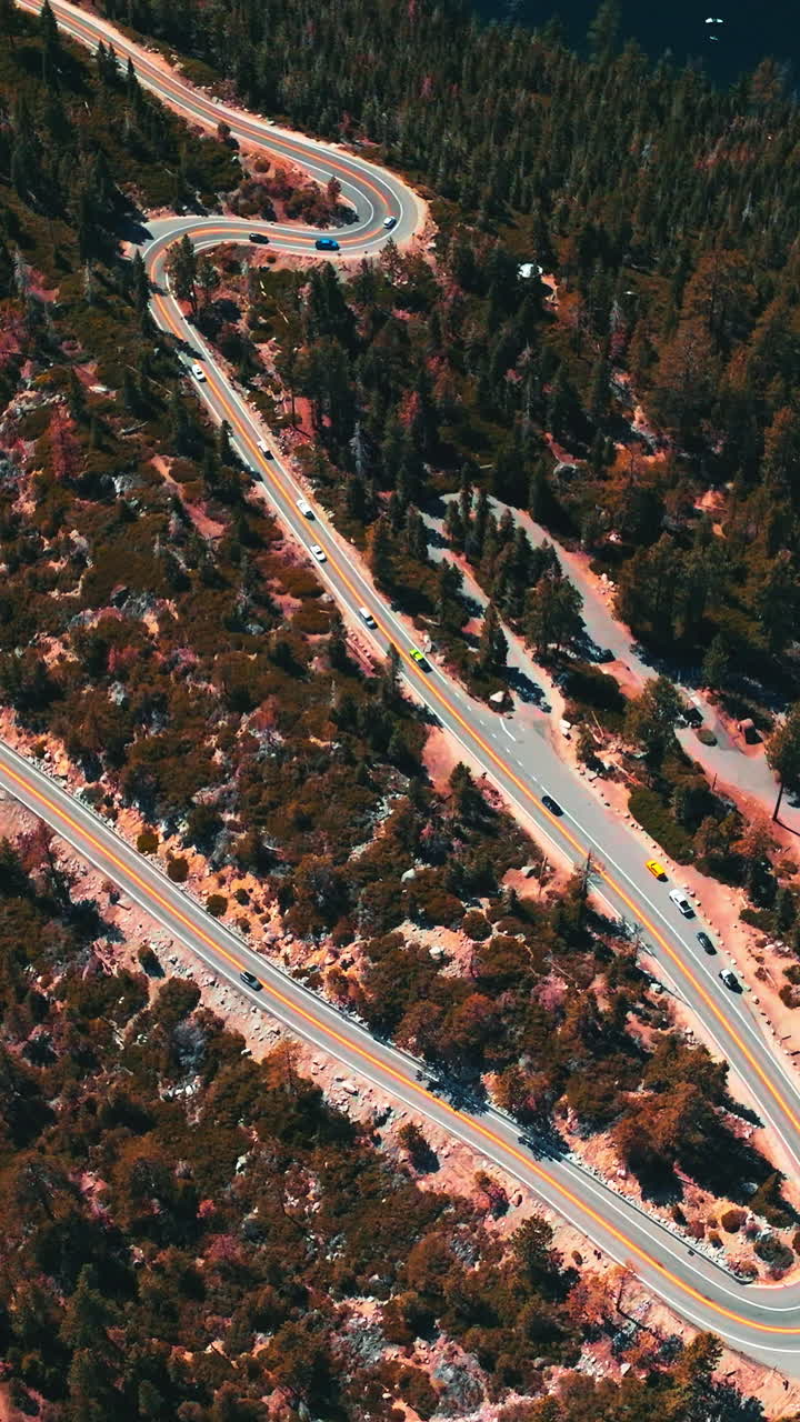 Twisted road through the wooded rocky landscape with many cars moving by. Deep blue lake at the foot of mountain from aerial perspective. Vertical video