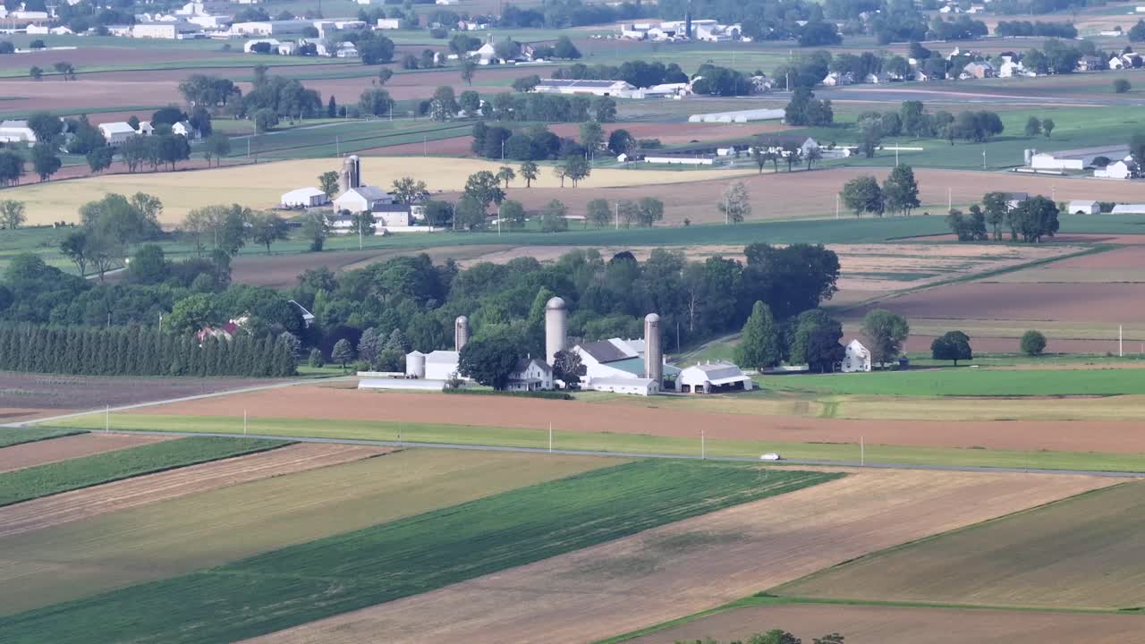 Car on rural road between farm fields with silo storages. Green and yellow cropland fields during sunny day in America. Descend drone wide shot.
