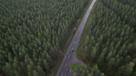 Vista-Aérea-De-Pájaro-De-La-Carretera-Forestal-En-Finlandia,-Coche-Negro-Conduciendo-Por-La-Carretera,-Dron-Siguiendo-El-Coche,-Tráfico,-Verano,-Día-Nublado
