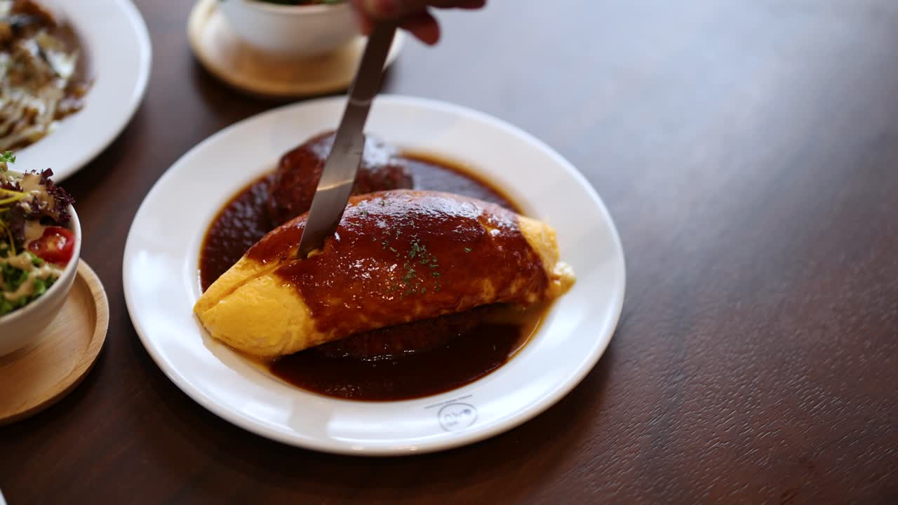 Hand slices fluffy Japanese omelette over rice, revealing creamy interior, warm lighting, close-up view