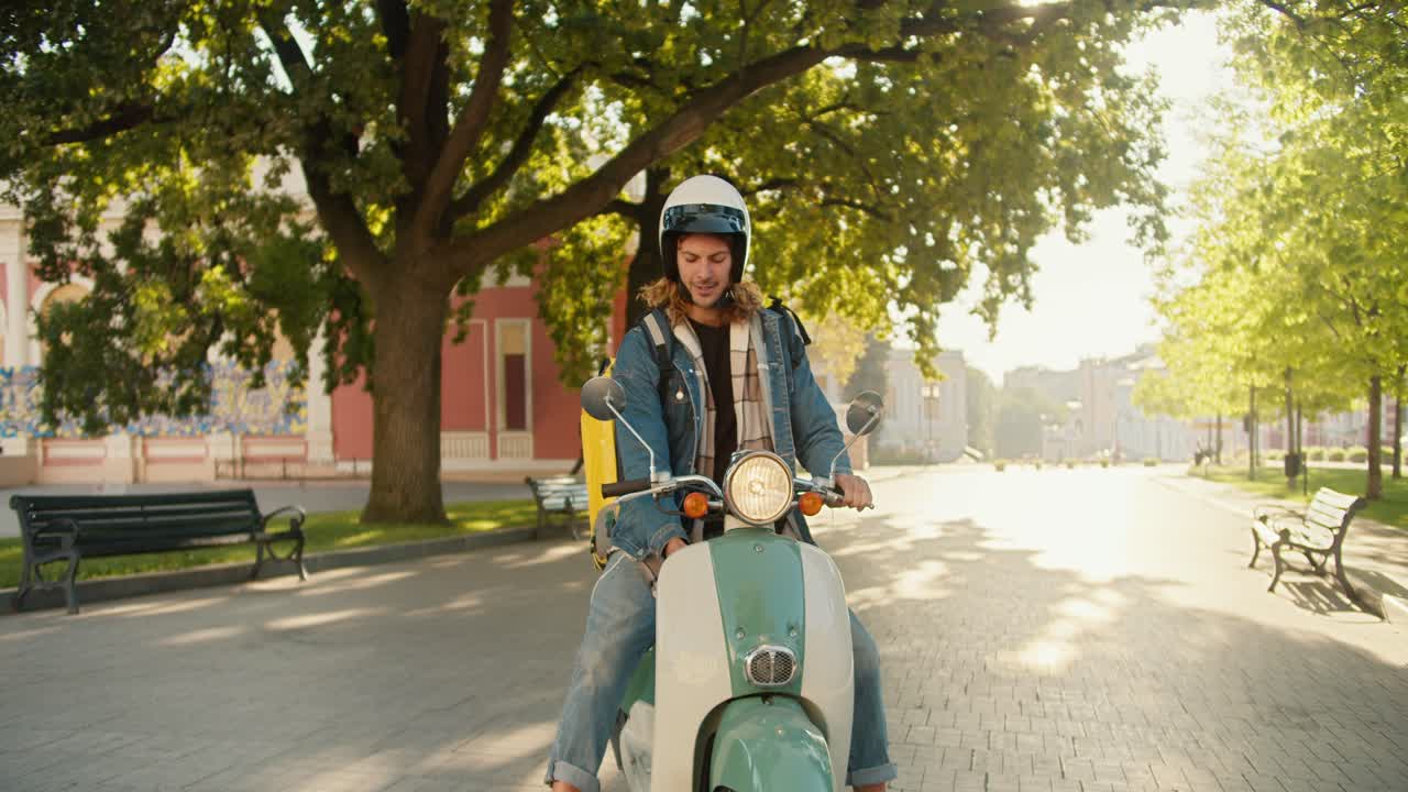 A happy courier guy with curly hair in a denim jacket arrived on a moped to a city park, looks around and looks on his phone to navigate where he should go next to deliver his order in a large yellow backpack