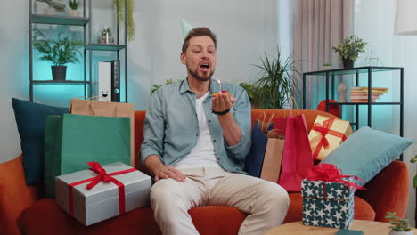 happy young man celebrating birthday party, makes wish blowing burning candle on small cupcake